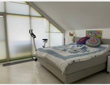 Zimmer unter einer Dachschräge mit großflächigen Fenstern, die mit Jalousien abgedeckt sind. Im Raum befinden sich ein graues Bett mit bunter Bettwäsche und runden Kissen, ein Heimtrainer sowie dekorative Elemente an der Wand.