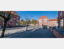 Straßenszene mit roten Backsteingebäuden im traditionellen Baustil, flankiert von Bäumen und Gehwegen. Im Hintergrund sind symmetrisch angeordnete Gebäude mit roten Dächern und weißen Fenstern sichtbar, bei klarem Himmel.