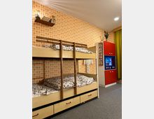 Etagenbett in einem Zimmer mit orange-gemusterter Tapete, Bettwäsche mit floralen Mustern, grauem Teppichboden und einem roten Schrank mit Fernseher. Oben links ist ein Regal mit Büchern angebracht, rechts hängt ein grüner Vorhang.