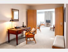 Helles Hotelzimmer mit einer Einrichtung aus Holz und Polstermöbeln. Im Vordergrund ein Schreibtisch mit Spiegel und Tischlampe, dahinter ein offener Wohnbereich mit Stühlen, einem Fernseher und großen Fenstern mit Vorhängen.