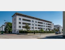 Mehrstöckiges Wohngebäude mit weißer Fassade und flachem Dach in einem Wohngebiet. Vor dem Gebäude befinden sich ein kleiner Gartenbereich mit Hecken, Gehwege, geparkte Autos und Straßenlaternen unter klarem Himmel.