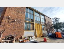 Backsteinfassade eines zweistöckigen Gebäudes mit großen, vergitterten Fenstern und einer verwitterten gelben Metalltür. Im Vordergrund liegen Baumaterialien und Abfall, daneben stehen Mülltonnen und ein Baum.