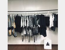 Kleiderstange mit unterschiedlich gemusterten Kleidungsstücken in Schwarz, Weiß und Grau vor einer weißen Wand; unten rechts steht eine weiße Einkaufstasche mit der Aufschrift „SEAM Berlin“ auf einem Holzboden.