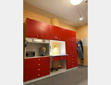 Ein Kinderzimmer im Retro-Stil mit einer Wand aus orange-gelb gemusterten Tapeten. Im Mittelpunkt steht eine rote Schrankwand mit weißen Knöpfen, die Schubladen, Regalfächer, einen Globus, ein Radio und einen integrierten Schreibtisch mit Schreibtischlampe enthält.