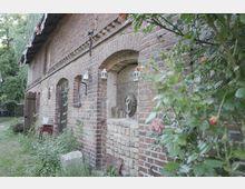 Backsteinmauer eines alten Gebäudes mit gewölbten Fenstern und Türen; an der Wand hängen kleine Laternen. Im Vordergrund befinden sich blühende Rosensträucher und ein kleiner Gartenbereich mit Pflanzen.
