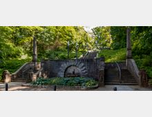 Steinerne Treppenanlage in einem parkähnlichen Gelände, umgeben von dichtem Grün und Bäumen, mit einem zentralen Brunnen an einer bogenförmigen Wand und symmetrisch angeordneten Stufen sowie steinernen Säulen.