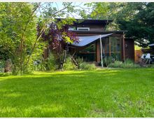 Einstöckiges Holzhaus mit großen Fenstern, umgeben von einem gepflegten Garten mit Rasen, Bäumen und Sträuchern. Im Vordergrund spannen Sonnensegel über der Terrasse, die teils von Pflanzen umgeben ist.