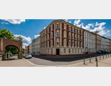 Straßenszene mit einer Ecke eines vierstöckigen Wohngebäudes im Altbaustil mit roter und weißer Fassade. Links ein gemauerter Torbogen, im Hintergrund Bäume und weiterer Wohnhäuser, unter klarem Himmel.