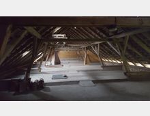 Dachboden eines Gebäudes mit offenen Holzträgern und sichtbarer Dachkonstruktion. Der Raum ist weitgehend leer, mit Betonboden und einzelnen Baumaterialien am Rand.