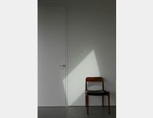 Weiße Wand mit einer schlichten Zimmertür links und einem hölzernen Stuhl mit schwarzer Sitzfläche rechts; ein schräger Lichteinfall bildet ein helles Dreieck auf der Wand.
