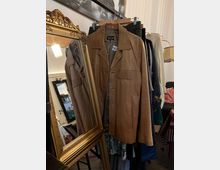 Ein brauner Ledermantel hängt an einem Kleiderbügel vor einem goldenen, verzierten Spiegel. Im Hintergrund sind weitere Kleidungsstücke auf einer Kleiderstange sowie Teile eines Raumes mit dekorativen Elementen erkennbar.