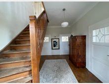 Holzgetäfelte Eingangshalle mit einer Treppe auf der linken Seite, die nach oben führt. Rechts ein antiker Holzschrank, eine helle Wand mit einem großen gerahmten Bild, und zwei Holztüren mit Milchglaseinsätzen.