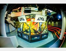 Ein moderner E-Sport-Veranstaltungsraum mit einem zentralen, verglasten Bühnenbereich in polygonaler Form, umgeben von Sitzreihen mit LED-Beleuchtung. Über der Bühne hängen Monitore mit dem Logo „LVL“, und die Umgebung ist mit technischen Elementen und Scheinwerfern ausgestattet.