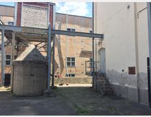 Ein Hof mit industriellem Charakter, umgeben von Gebäuden mit verputzten Fassaden und freiliegenden Metallgerüsten. Im Vordergrund steht eine kleine Betonstruktur mit spitzem Dach, daneben eine Metalltreppe, die zu einer Tür führt.