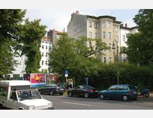 Straßenszene mit mehrstöckigen Wohnhäusern im Hintergrund, umgeben von Bäumen und Grünflächen. Im Vordergrund parken Autos entlang der Straße, und mehrere bunte Werbetafeln sind zu sehen.