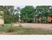 Grüne Außenanlage mit einem Sand-Volleyballfeld in der Mitte, umgeben von Bäumen und Wiesen. Im Hintergrund befinden sich gelb verputzte Gebäude mit roten Dächern und ein kleiner Spielplatz mit Schaukeln und einer Rutsche.