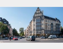 Straßenkreuzung in einer Stadt mit einem historischen, mehrstöckigen Gebäude im Jugendstil als zentralem Motiv. Das helle Gebäude hat dekorative Fassadenelemente, große Fenster und ein spitzes Dach, umgeben von Bäumen, weiteren Wohnhäusern und vorbeifahrenden Autos.