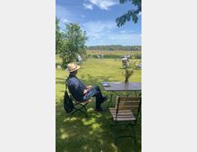 Ein Garten mit Rasenfläche und verstreut stehenden Tischen und Stühlen, an denen Menschen entspannt sitzen. Im Hintergrund befinden sich Bäume und ein Teich unter einem leicht bewölkten blauen Himmel.