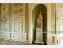 Klassischer Innenraum mit einer weißen Doppeltür, die von pastellfarbenen Wanddekorationen mit floralen und antiken Motiven umgeben ist. Eine Marmorskulptur in einer grün akzentuierten Nische steht auf einem verzierten Sockel, der an den Stil des Raumes angepasst ist.