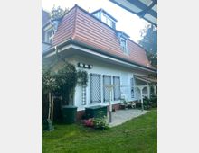 Ein Einfamilienhaus mit rot gedecktem Dach, großen Dachfenstern und weißer Fassade. Der Garten zeigt einen Rasen, Blumenbeete, Rankgitter an der Wand und eine Terrasse mit Pflastersteinen, Stühlen und einer Grünfläche.