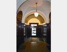 Eingangsbereich eines historischen Gebäudes mit Holzwänden, verziertem Rundbogen und einem zentral hängenden Lampenschirm. Eine verglaste Tür mit Blick auf die Straße bildet den Ausgang.