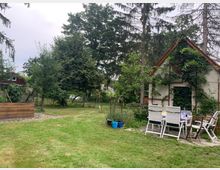 Ein Garten mit einer Wiese, umgeben von Bäumen und Büschen. Rechts steht ein kleines Haus mit Kletterpflanzen an der Fassade, davor ein Gartentisch mit weißen Stühlen.