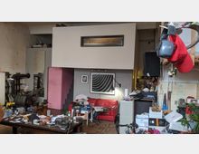 Industrieähnlicher Raum mit einer erhöhten Zwischendecke und rechteckigem Fenster, darunter eine Wand mit moderner Kunst und einem roten Ledersofa. Der Raum ist unaufgeräumt, mit Arbeitsmaterialien, Werkzeugen und Möbeln verstreut, sowie Regalen und Dekoration an den Wänden.