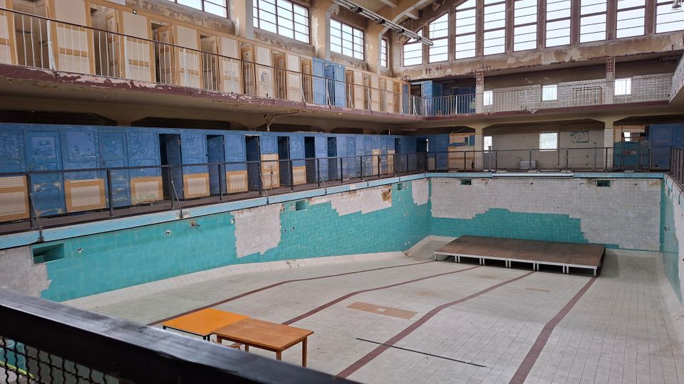 Ehemaliges Schwimmbad mit leeren, trockenen Becken, türkisfarbenen Fliesen und teils abgebröckelten Wänden. Im oberen Bereich sind Umkleidekabinen und Geländer sichtbar, begleitet von großen Fenstern, die natürliches Licht hereinlassen.