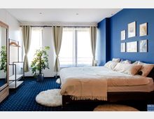 Schlafzimmer mit einem großen Bett, blauer Akzentwand und mehreren gerahmten Bildern. Helle Vorhänge an drei Fenstern, ein Teppich mit geometrischem Muster, ein Kleiderständer aus Holz und Metall sowie eine Zimmerpflanze ergänzen die Einrichtung.