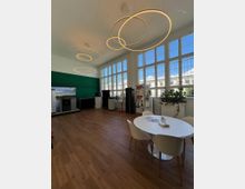 Ein moderner Showroom mit hohen Fenstern, die viel Tageslicht einlassen, einer weißen Decke mit kreisförmigen, leuchtenden Hängeleuchten, einem Holzboden, und einem weißen runden Tisch mit Stühlen im Vordergrund. Im Hintergrund stehen verschiedene Geräte und Ausstellungsstücke vor einer dunkelgrünen Wand.