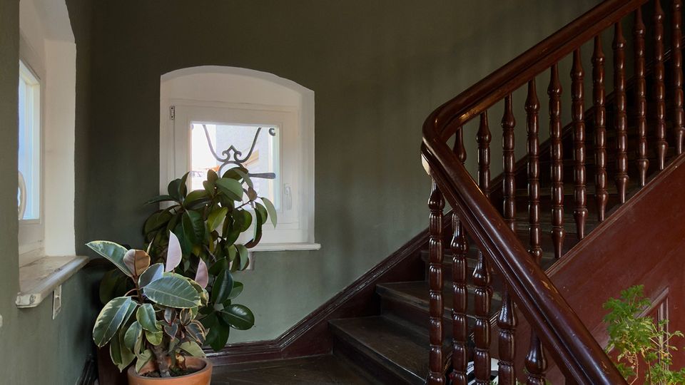 Innenansicht eines Treppenhauses mit dunkler Holztreppe und gedrechseltem Geländer, grün gestrichenen Wänden und weißer Decke. Auf dem Treppenabsatz stehen Topfpflanzen neben einem kleinen Fenster mit weißem Rahmen.