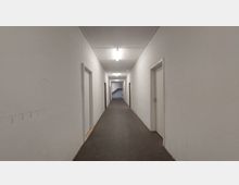 Langer, schlichter Flur mit weiß gestrichenen Wänden, dunklem Bodenbelag und mehreren geschlossenen weißen Türen auf beiden Seiten. Am Ende des Flurs führen Treppen nach oben, und die Decke ist mit einfachen Deckenlampen beleuchtet.