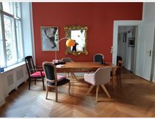 Esszimmer mit roten Wänden, einem Holztisch und verschiedenen Stühlen, darunter ein rosafarbener und ein gepolsterter. An der Wand hängt ein goldgerahmter Spiegel und ein gerahmtes Bild. Links sind große Fenster mit Blick ins Freie sichtbar.