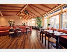 Bright restaurant dining room with wood floors, red upholstered banquettes, and dark tables set with plates and wine glasses. A high vaulted ceiling with exposed beams and hanging lantern lights sits above large glass doors and windows overlooking trees, with potted plants and cacti as decor.