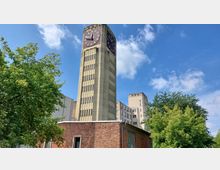 Gelber Uhrturm mit großer analoger Uhr vor einem blauen Himmel, daneben niedrigere Backsteingebäude und moderne Bürogebäude im Hintergrund; im Vordergrund sind Bäume und Grünflächen zu sehen.