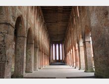 Innenansicht einer gotischen Backsteinkirche mit hohen Spitzbögen, massiven Säulen und einem offenen Dachstuhl. Am Ende des langen Kirchenschiffs sind große Spitzbogenfenster mit farblosem Glas zu sehen.
