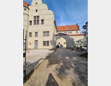 Historisches Gebäude mit hellbeiger Fassade und roten Ziegeldächern, im Stil mit geschwungenem Giebel. Ein offener Torbogen führt zu einem Innenhof; der Boden davor ist teils gepflastert, teils mit Sand bedeckt, und Bauarbeiten sind erkennbar.