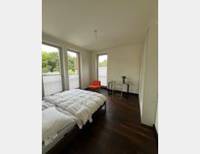 Helles Schlafzimmer mit dunklem Holzboden, einem Doppelbett mit weißer Bettwäsche, drei großen Fenstern mit Blick ins Grüne und einem Eckbereich mit einem kleinen Schreibtisch, einem Stuhl und einem orangefarbenen Sessel. Ein großer, weißer Kleiderschrank steht rechts an der Wand.