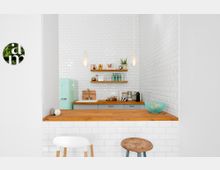 Minimalistische Küche mit weißen Fliesenwänden, einer Holzarbeitsplatte und offenen Holzregalen mit Geschirr und Dekoration. Ein mintgrüner Kühlschrank, eine Spüle sowie eine Kaffeemaschine sind sichtbar; im Vordergrund steht eine Theke mit Hockern.