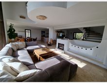 Wohnzimmer in einem modernen Haus mit offenem Grundriss. Zu sehen sind eine große Eckcouch, ein Holztisch, ein offener Kamin mit weißem Putz und runden Verzierungen, eine Holztreppe sowie ein Essbereich mit Tisch und Stühlen im Hintergrund.