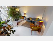 Helles Wohnzimmer mit weißem Boden, einer grauen Eckcouch mit bunten Kissen, einem kleinen Holztisch und einem Sessel mit gelbem Kissen. Im Raum befinden sich mehrere Pflanzen, eine hängende Lampe und ein Sideboard mit Lautsprechern und Dekoration, beleuchtet durch warme Lampen.