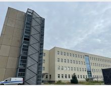 Außenansicht eines mehrstöckigen, hell verputzten Büro- oder Verwaltungsgebäudes mit vielen gleichmäßig angeordneten Fenstern und einem verglasten Mittelteil. Links ist ein angebauter, hoher Treppen- oder Aufzugsturm aus Metall und Glas mit perforierten Paneelen und diagonalen Streben zu sehen, davor ein kleiner Vorplatz mit etwas Grünfläche.