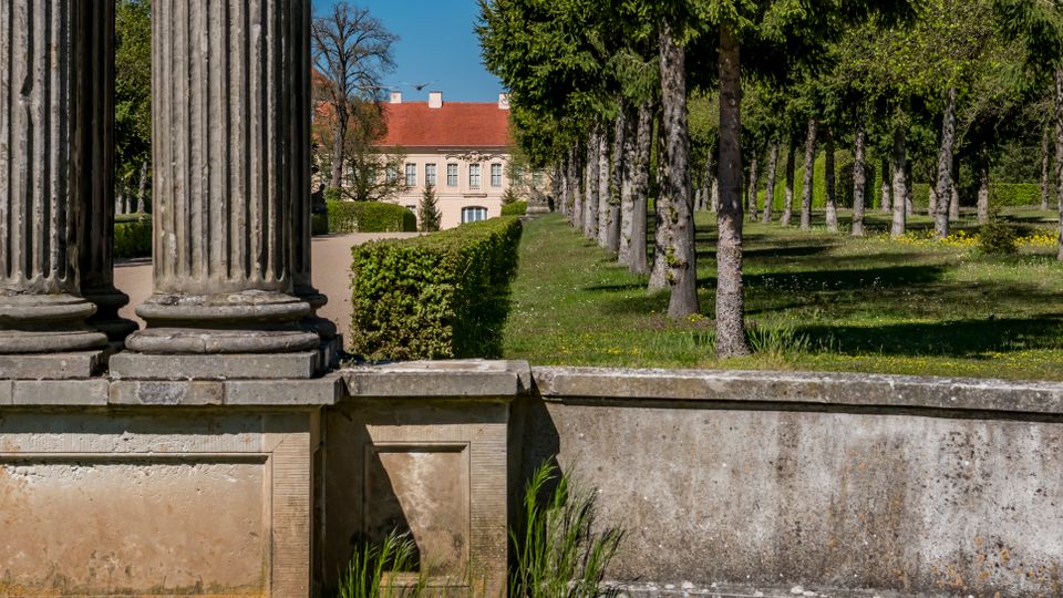 View from a stone terrace with large fluted classical columns, looking along a manicured garden with clipped hedges and rows of trees. A pale historic manor with a red-tiled roof stands in the distance beyond the formal grounds.