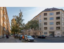 Eine straßenansicht in einer Wohngegend mit gelblichen Mehrfamilienhäusern im Bauhausstil, Bäumen entlang des Gehwegs und geparkten Autos auf der Straße. Ein Straßenschild mit der Aufschrift „Herschelstraße“ steht an einer Kreuzung, und im Erdgeschoss ist die Beschilderung einer „Yoga Schule“ erkennbar.