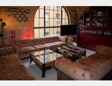 Ein stilvoll eingerichteter Raum mit Backsteinwänden, großen Bogenfenstern und braunen Ledersofas in Chesterfield-Stil. Im Hintergrund befindet sich ein rotes Holzregal mit Flaschen und Dekorartikeln, daneben ein Fernseher auf einer Holzpalette.