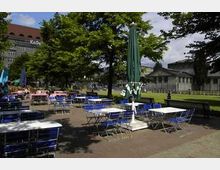 Außenbereich eines Cafés mit Tischen und blau-weißen Stühlen auf einem gepflasterten Platz, umgeben von Bäumen. Im Hintergrund sind historische Gebäude und Grünflächen zu sehen, teils im Schatten der Baumkronen.