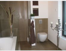 Modernes Badezimmer mit beige-braunen Fliesen, einer weißen Wandtoilette und einer Badewanne links im Bild. Dekorationselemente umfassen eine Vase mit Zweigen, ein Handtuchhalter mit braunem Handtuch, eine Nische mit Handtüchern, eine kleine Pflanze und einen silbernen Kerzenständer mit mehreren Armen.