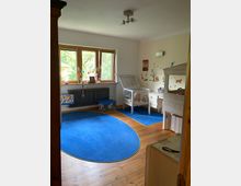 Ein Kinderzimmer mit Holzboden, einem großen, runden, blauen Teppich und einem hölzernen Etagenbett auf der rechten Seite. An der Wand links stehen ein Fenster mit Holzrahmen, ein Heizkörper und ein kleiner Tisch mit Dekoration; an der rechten Wand befinden sich kindgerechte Wandsticker sowie ein Kleintiergehege mit transparentem Aufsatz.