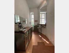 Schmale Küche mit weißen Wänden und hohen Fenstern, ausgestattet mit einem langen Tresen mit Kaffeemaschine und Glaskühlschränken darunter. Rechts befinden sich ein Hocker und eine schmale Ablage an der Wand, durch die Fenster fällt natürliches Licht.