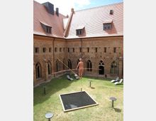 Innenhof eines historischen Backsteingebäudes mit gotischen Spitzbogenfenstern, umgeben von Rasen. In der Mitte balanciert eine Figur auf einem Drahtseil über einem rechteckigen Wasserbecken.